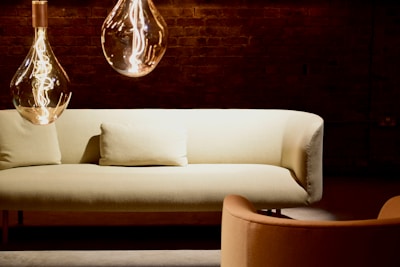 A cozy and modern living space featuring a light-colored sofa with a matching pillow in front of a dark brick wall. Two large, stylish pendant lights with visible filaments hang from the ceiling, casting a warm glow. An armchair in complementary tones is situated nearby.