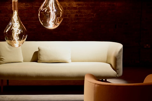 A cozy and modern living space featuring a light-colored sofa with a matching pillow in front of a dark brick wall. Two large, stylish pendant lights with visible filaments hang from the ceiling, casting a warm glow. An armchair in complementary tones is situated nearby.