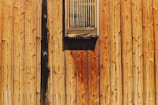 A wooden wall with vertical planks, featuring a small, rectangular, slatted window. The wood displays a rich grain pattern with shades of brown and a slightly rustic appearance.
