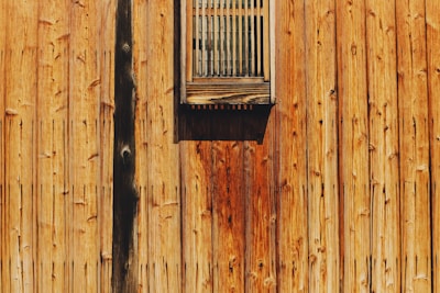 A wooden wall with vertical planks, featuring a small, rectangular, slatted window. The wood displays a rich grain pattern with shades of brown and a slightly rustic appearance.