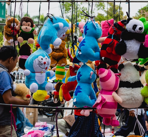 A bustling street market scene features a variety of colorful plush toys hanging for sale. The toys include characters such as pandas, blue tigers, monkeys, a frog, and others in vibrant colors like blue, pink, red, and green. A man on the left side engages with the display, creating a lively and playful atmosphere.