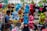 A bustling street market scene features a variety of colorful plush toys hanging for sale. The toys include characters such as pandas, blue tigers, monkeys, a frog, and others in vibrant colors like blue, pink, red, and green. A man on the left side engages with the display, creating a lively and playful atmosphere.