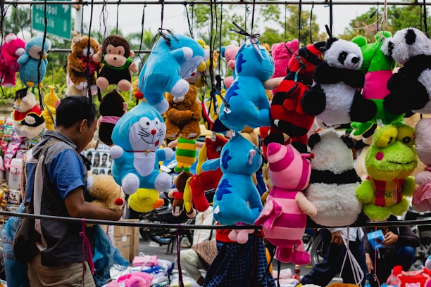 A bustling street market scene features a variety of colorful plush toys hanging for sale. The toys include characters such as pandas, blue tigers, monkeys, a frog, and others in vibrant colors like blue, pink, red, and green. A man on the left side engages with the display, creating a lively and playful atmosphere.