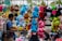A bustling street market scene features a variety of colorful plush toys hanging for sale. The toys include characters such as pandas, blue tigers, monkeys, a frog, and others in vibrant colors like blue, pink, red, and green. A man on the left side engages with the display, creating a lively and playful atmosphere.