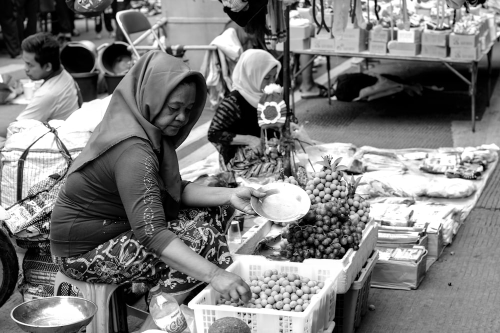 Photographie en niveaux de gris d’une femme assise à côté de fruits dans un panier