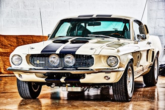 A vintage muscle car is parked indoors on a shiny wooden floor. The car has a cream-colored body with prominent black racing stripes running from the front hood to the roof. The headlights are circular, and there are three round fog lights integrated into the front grille. The car's exterior is highly polished and reflective, and it has a sleek, aerodynamic design with large, visible tires and a slightly aggressive stance.