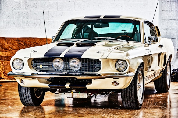 A vintage muscle car is parked indoors on a shiny wooden floor. The car has a cream-colored body with prominent black racing stripes running from the front hood to the roof. The headlights are circular, and there are three round fog lights integrated into the front grille. The car's exterior is highly polished and reflective, and it has a sleek, aerodynamic design with large, visible tires and a slightly aggressive stance.