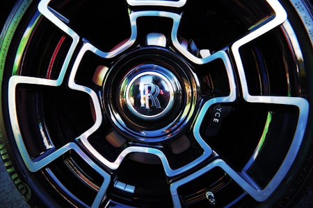 A close-up view of a car wheel featuring a distinctive and intricate alloy design with a shiny, reflective surface. The center cap prominently displays the letter 'R' within a polished emblem, surrounded by a symmetrical pattern that combines sleek black and metallic silver elements.