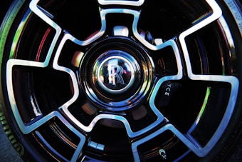 A close-up view of a car wheel featuring a distinctive and intricate alloy design with a shiny, reflective surface. The center cap prominently displays the letter 'R' within a polished emblem, surrounded by a symmetrical pattern that combines sleek black and metallic silver elements.