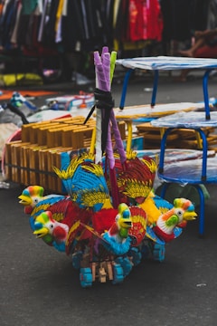 Vibrant, multicolored toy parrots made of wood and fabric are clustered together on the ground. The toys are detailed with red, blue, yellow, and green, showcasing intricate feather designs. In the background, a busy street market setting unfolds, with tables, racks, and various items for sale visible.
