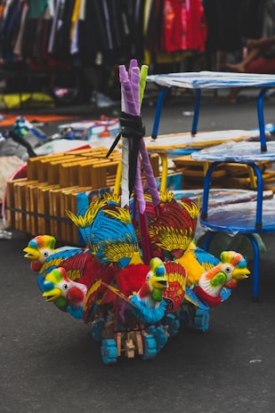 Vibrant, multicolored toy parrots made of wood and fabric are clustered together on the ground. The toys are detailed with red, blue, yellow, and green, showcasing intricate feather designs. In the background, a busy street market setting unfolds, with tables, racks, and various items for sale visible.