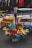 Vibrant, multicolored toy parrots made of wood and fabric are clustered together on the ground. The toys are detailed with red, blue, yellow, and green, showcasing intricate feather designs. In the background, a busy street market setting unfolds, with tables, racks, and various items for sale visible.