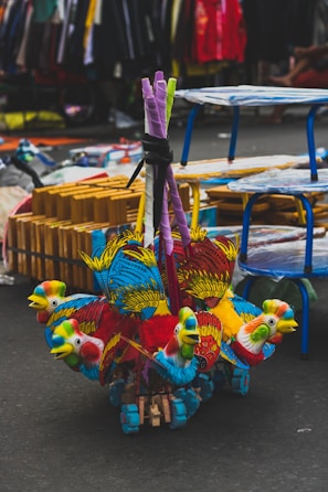 Vibrant, multicolored toy parrots made of wood and fabric are clustered together on the ground. The toys are detailed with red, blue, yellow, and green, showcasing intricate feather designs. In the background, a busy street market setting unfolds, with tables, racks, and various items for sale visible.