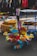 Vibrant, multicolored toy parrots made of wood and fabric are clustered together on the ground. The toys are detailed with red, blue, yellow, and green, showcasing intricate feather designs. In the background, a busy street market setting unfolds, with tables, racks, and various items for sale visible.