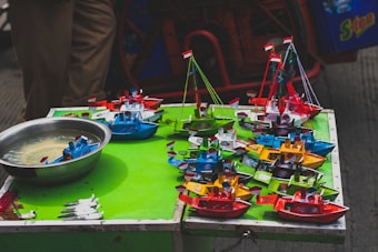 A collection of colorful miniature toy boats displayed on a vibrant green surface. Each boat is uniquely colored in bright shades such as red, blue, yellow, and green, featuring small flags and intricate details. A metal bowl with water holds some of the boats, likely demonstrating their buoyancy. The scene suggests a lively street market setup with a casual arrangement.