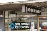 A storefront features a sign for a record shop called 'Goldmine Records,' with a visible logo consisting of a stylized, blue and black vinyl record. The shop window displays various music-related posters and advertisements, while the entrance door has store hours and more promotional material. The atmosphere suggests a vintage or retro theme.