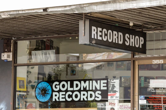 A storefront features a sign for a record shop called 'Goldmine Records,' with a visible logo consisting of a stylized, blue and black vinyl record. The shop window displays various music-related posters and advertisements, while the entrance door has store hours and more promotional material. The atmosphere suggests a vintage or retro theme.