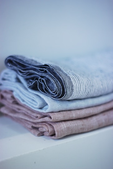 A neatly folded stack of linens with a soft, textured appearance. The linens are in muted tones, arranged on a white surface with a soft background blur.