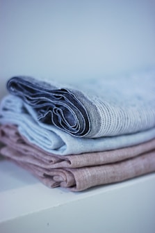 A neatly folded stack of linens with a soft, textured appearance. The linens are in muted tones, arranged on a white surface with a soft background blur.