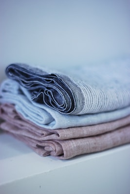 A neatly folded stack of linens with a soft, textured appearance. The linens are in muted tones, arranged on a white surface with a soft background blur.
