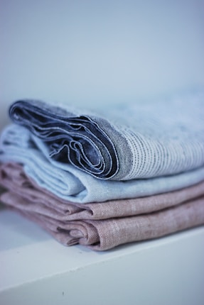A neatly folded stack of linens with a soft, textured appearance. The linens are in muted tones, arranged on a white surface with a soft background blur.