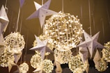 Hanging decorations feature illuminated wire spheres intertwined with string lights and paper stars adorned with small stars, creating a festive and whimsical atmosphere.
