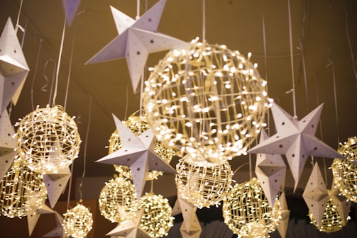 Hanging decorations feature illuminated wire spheres intertwined with string lights and paper stars adorned with small stars, creating a festive and whimsical atmosphere.