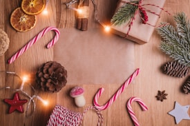 A festive arrangement on a wooden surface featuring wrapped gifts with pine branches, candy canes, dried orange slices, cinnamon sticks, and other holiday decorations such as pine cones and stars. Twinkling lights add warmth to the setup.