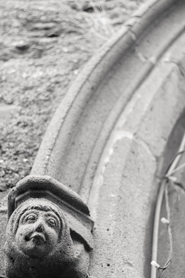 A stone carving of a face with a serene expression is integrated into a curved architectural structure. The texture of the stone is rough, with visible wear over time. The background includes a blurred textured surface, potentially gravel.
