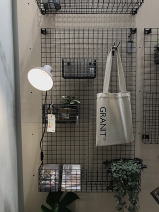 A metal grid wall organizer with shelves and baskets holds various objects. A hanging beige tote bag reads 'GRANIT'. A small potted plant is set on one of the shelves, and a wall-mounted reading lamp with a white shade illuminates the area. Printed materials lie on the bottom shelf with a green leafy plant next to them.