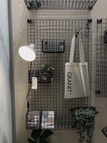 A metal grid wall organizer with shelves and baskets holds various objects. A hanging beige tote bag reads 'GRANIT'. A small potted plant is set on one of the shelves, and a wall-mounted reading lamp with a white shade illuminates the area. Printed materials lie on the bottom shelf with a green leafy plant next to them.