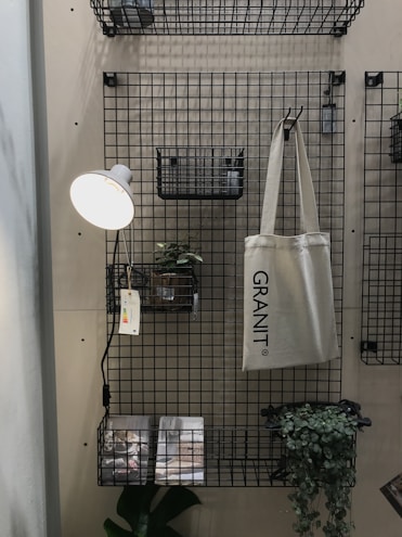 A metal grid wall organizer with shelves and baskets holds various objects. A hanging beige tote bag reads 'GRANIT'. A small potted plant is set on one of the shelves, and a wall-mounted reading lamp with a white shade illuminates the area. Printed materials lie on the bottom shelf with a green leafy plant next to them.