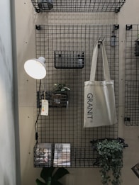 A metal grid wall organizer with shelves and baskets holds various objects. A hanging beige tote bag reads 'GRANIT'. A small potted plant is set on one of the shelves, and a wall-mounted reading lamp with a white shade illuminates the area. Printed materials lie on the bottom shelf with a green leafy plant next to them.