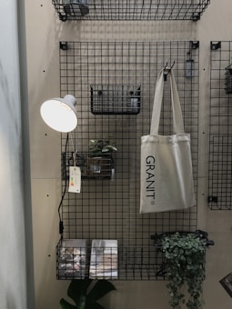 A metal grid wall organizer with shelves and baskets holds various objects. A hanging beige tote bag reads 'GRANIT'. A small potted plant is set on one of the shelves, and a wall-mounted reading lamp with a white shade illuminates the area. Printed materials lie on the bottom shelf with a green leafy plant next to them.