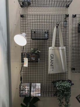 A metal grid wall organizer with shelves and baskets holds various objects. A hanging beige tote bag reads 'GRANIT'. A small potted plant is set on one of the shelves, and a wall-mounted reading lamp with a white shade illuminates the area. Printed materials lie on the bottom shelf with a green leafy plant next to them.