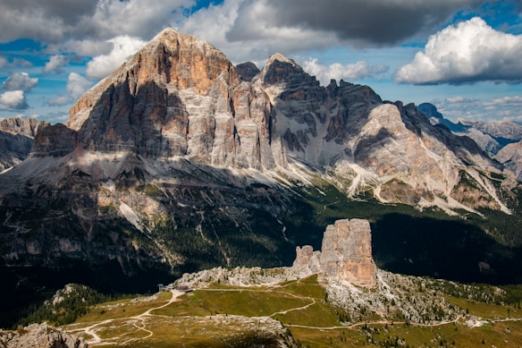 A stunning mountainous landscape featuring a large, imposing rock formation with various shades of brown and gray. The scene includes a smaller rock formation in the foreground, surrounded by lush green vegetation and intricate hiking trails. The sky is partially cloudy with a mix of white and dark clouds.