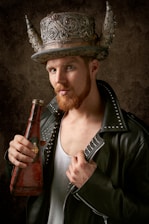 A man with a beard wearing a leather jacket adorned with studs is holding an ornate, weathered bottle. He sports a large, decorative hat featuring intricate designs and prominent horns. The background is a textured, earthy tone.
