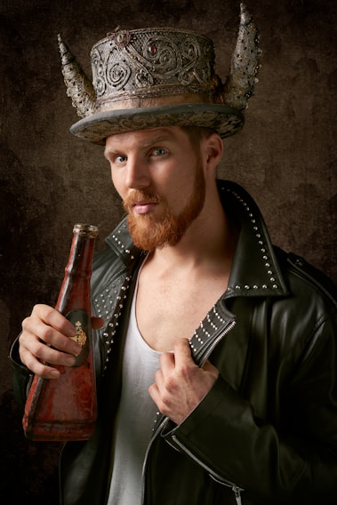 A man with a beard wearing a leather jacket adorned with studs is holding an ornate, weathered bottle. He sports a large, decorative hat featuring intricate designs and prominent horns. The background is a textured, earthy tone.