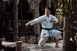 A martial artist in white traditional attire performs a poised karate stance on a wooden platform in a forested area. The sunlight filters through the trees, casting dappled light across the scene.