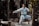 A martial artist in white traditional attire performs a poised karate stance on a wooden platform in a forested area. The sunlight filters through the trees, casting dappled light across the scene.