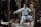 A martial artist in white traditional attire performs a poised karate stance on a wooden platform in a forested area. The sunlight filters through the trees, casting dappled light across the scene.