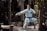A martial artist in white traditional attire performs a poised karate stance on a wooden platform in a forested area. The sunlight filters through the trees, casting dappled light across the scene.