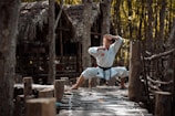 A person dressed in a white martial arts uniform stands in a wide stance with hands in a defensive position on a rustic wooden path in a forest setting. The background consists of trees and a thatched-roof structure, suggesting a natural or rural environment.