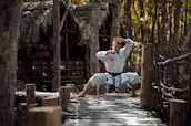A person dressed in a white martial arts uniform stands in a wide stance with hands in a defensive position on a rustic wooden path in a forest setting. The background consists of trees and a thatched-roof structure, suggesting a natural or rural environment.