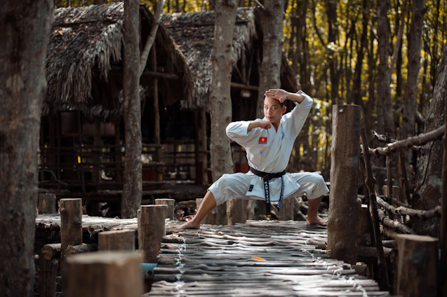 A person dressed in a white martial arts uniform stands in a wide stance with hands in a defensive position on a rustic wooden path in a forest setting. The background consists of trees and a thatched-roof structure, suggesting a natural or rural environment.