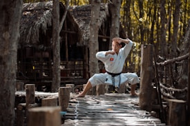 A person dressed in a white martial arts uniform stands in a wide stance with hands in a defensive position on a rustic wooden path in a forest setting. The background consists of trees and a thatched-roof structure, suggesting a natural or rural environment.