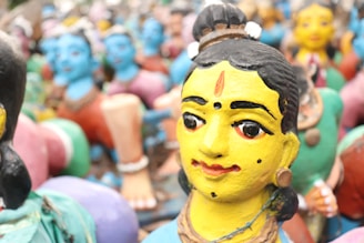 A group of colorful, traditional clay figurines depicting human figures in vibrant attire. The figures have painted faces with distinct expressions and intricate details. The foreground shows a close-up of one figurine with a yellow face and black hair, adorned with a bindi and earrings.