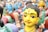 A group of colorful, traditional clay figurines depicting human figures in vibrant attire. The figures have painted faces with distinct expressions and intricate details. The foreground shows a close-up of one figurine with a yellow face and black hair, adorned with a bindi and earrings.