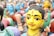 A group of colorful, traditional clay figurines depicting human figures in vibrant attire. The figures have painted faces with distinct expressions and intricate details. The foreground shows a close-up of one figurine with a yellow face and black hair, adorned with a bindi and earrings.