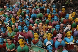 A large group of colorful, handcrafted clay figurines representing women with various poses and expressions. The figurines feature bright blue and yellow faces, each adorned with unique hairstyles and traditional attire in shades of red, green, and blue. They are closely packed together, creating a vibrant and diverse display of artistic craftsmanship.